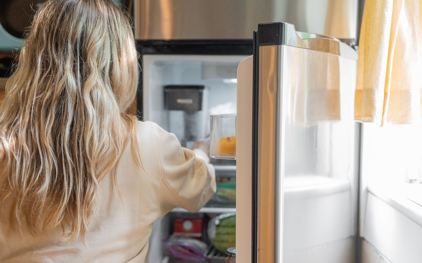 A woman standing at an open refrigerator, reaching inside, seen from behind